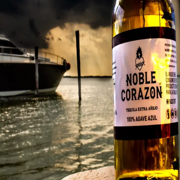 Close-up of a premium Extra Añejo 100% Agave Azul tequila bottle with a luxury yacht and dark, dramatic sky in the background, promoting a high-end experience in Cancun.